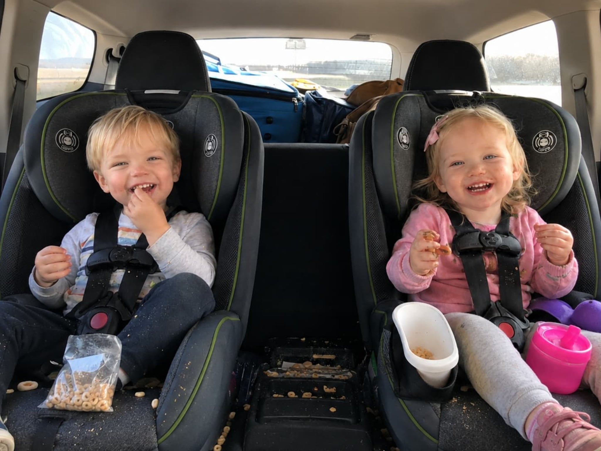 Two children sitting in car seats with snacks and a pink cup in the backseat of a car.