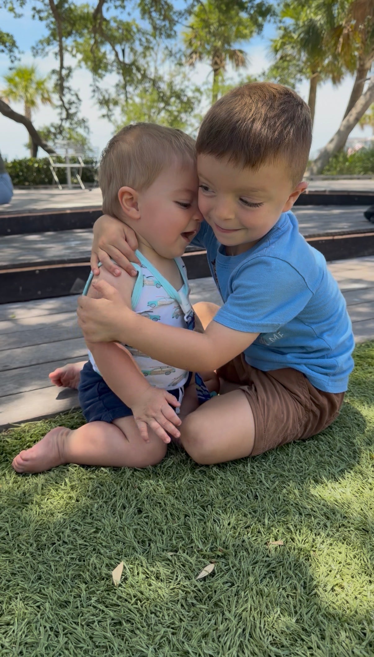 Two young boys sitting on grass, hugging each other.