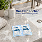 A Bubbez WASH and RINSE twin sachet lies open on a marble bathroom vanity beside the sink, a folded towel and a plant. Headline reads One twin sachet. Washed. Rinsed. No sticky residue.