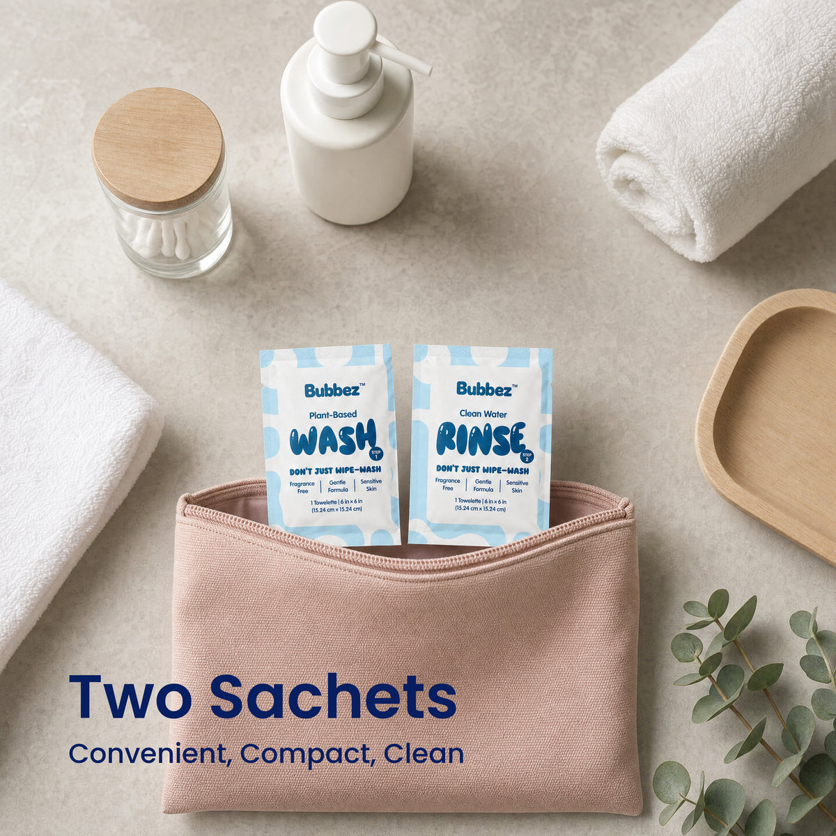 A Bubbez WASH and RINSE twin sachet tucked into a pink cosmetic pouch on a stone vanity with rolled towels, a pump bottle, cotton buds and eucalyptus. Headline reads Two Sachets. Convenient, Compact, Clean.