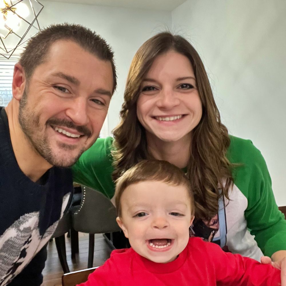 Family of three smiling indoors, likely in a home setting.