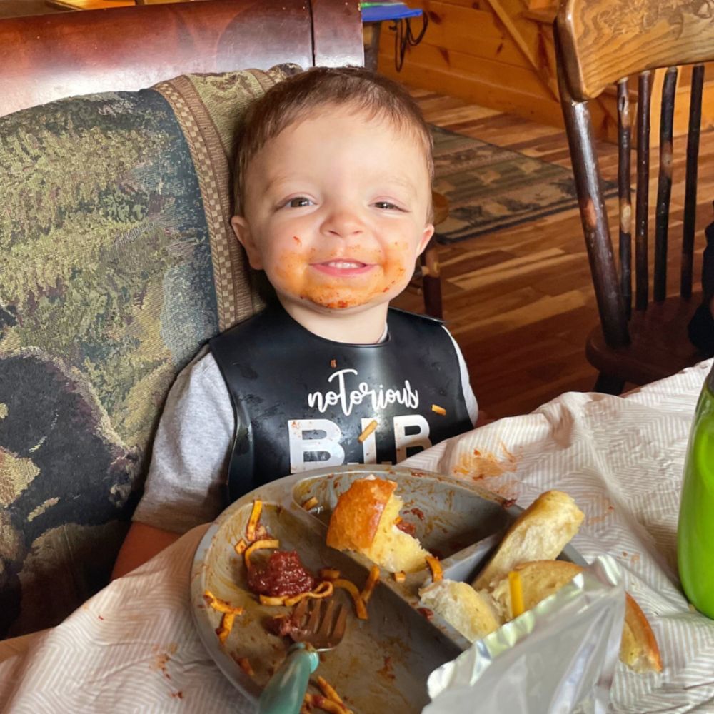 Child with food on face and shirt, sitting at a table with a plate of food.