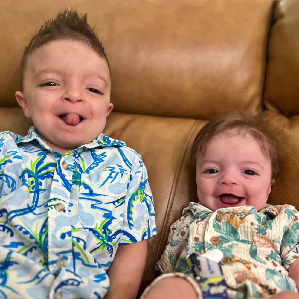Two children sitting on a brown couch, one sticking out their tongue and the other smiling.