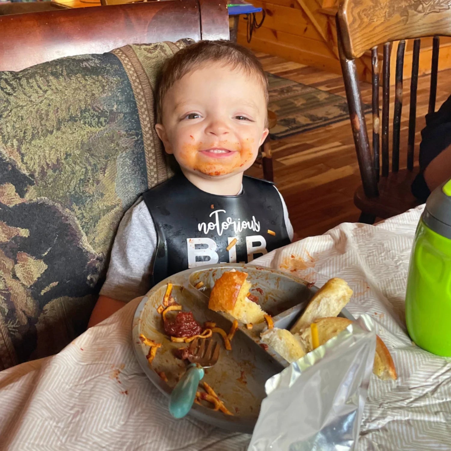 Child with food on face and shirt, sitting at a table with a plate of food.