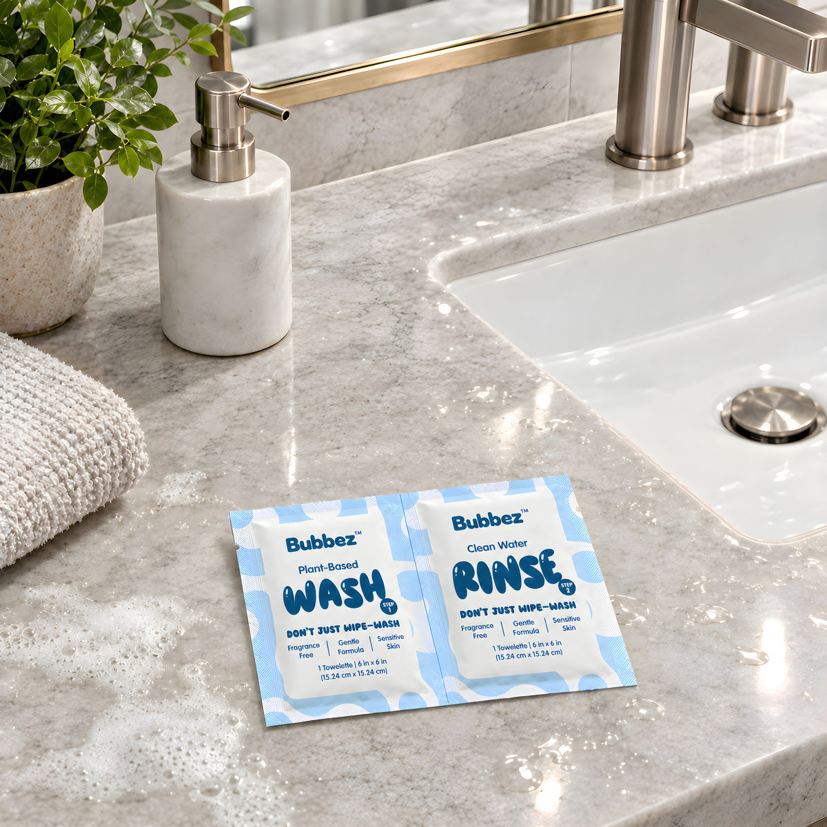 A Bubbez WASH and RINSE twin sachet lies open on a marble bathroom vanity beside the sink, a soap pump, a folded towel and a plant, with suds on the counter.