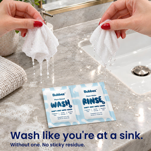 Two hands hold a lathering Bubbez WASH wipe and a dripping RINSE wipe above a gray marble countertop with a sink and a towel in frame. A WASH and RINSE twin sachet lies on the counter below.